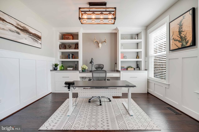 office featuring a decorative wall, healthy amount of natural light, built in shelves, dark wood-style flooring, and wainscoting