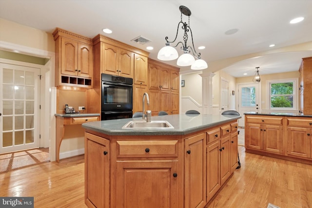 kitchen with a sink, arched walkways, light wood finished floors, decorative columns, and an island with sink