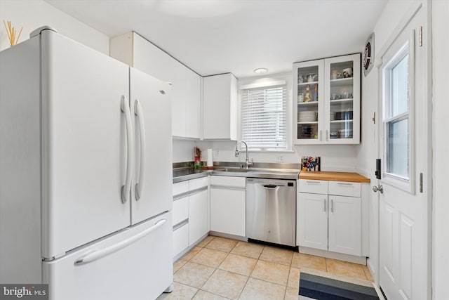 kitchen featuring freestanding refrigerator, stainless steel dishwasher, light tile patterned floors, white cabinets, and glass insert cabinets