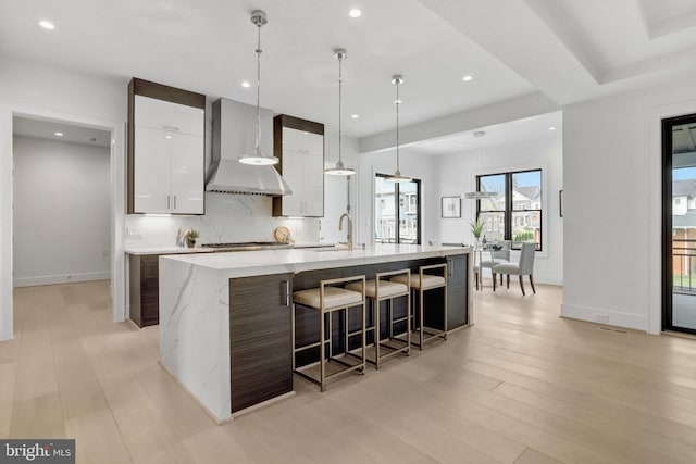 kitchen with wall chimney exhaust hood, modern cabinets, white cabinets, a large island with sink, and backsplash