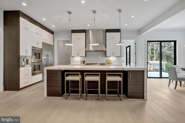 kitchen with wall chimney range hood, modern cabinets, light wood-style floors, tasteful backsplash, and stainless steel appliances