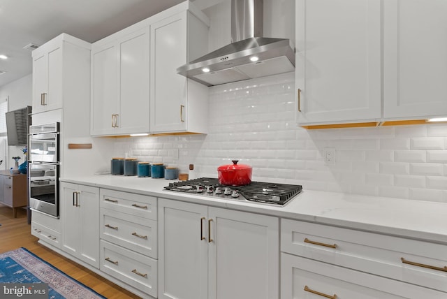 kitchen featuring wall chimney range hood, white cabinets, light wood-style flooring, appliances with stainless steel finishes, and backsplash