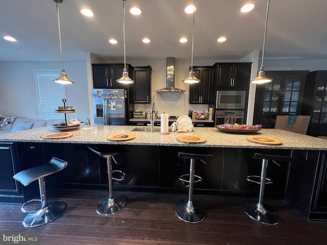 kitchen with appliances with stainless steel finishes, wall chimney exhaust hood, a large island, dark wood-style flooring, and a kitchen bar