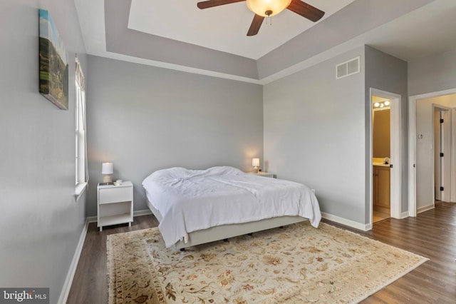 bedroom featuring wood finished floors, baseboards, a tray ceiling, ceiling fan, and ensuite bathroom