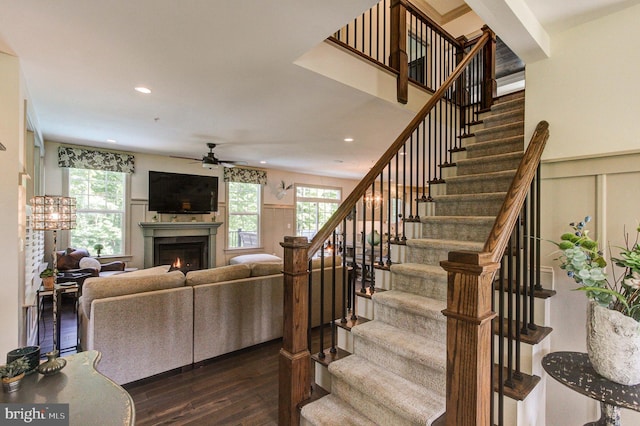 living room with ceiling fan, dark wood-style floors, stairs, a lit fireplace, and a decorative wall