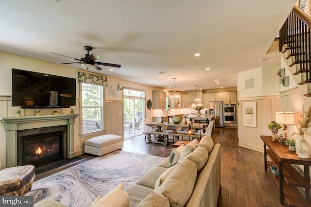 living area featuring a decorative wall, dark wood-type flooring, a ceiling fan, a fireplace with flush hearth, and a wainscoted wall