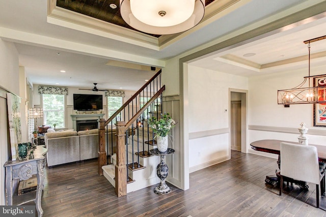 stairs featuring a tray ceiling, a chandelier, a ceiling fan, a lit fireplace, and crown molding