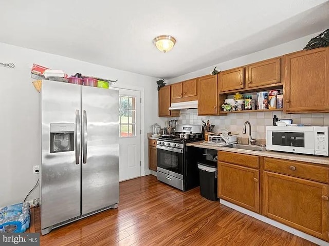 kitchen with stainless steel appliances, a sink, under cabinet range hood, brown cabinets, and dark wood-type flooring