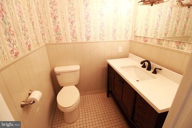 half bathroom with vanity, toilet, wallpapered walls, a wainscoted wall, and tile patterned floors