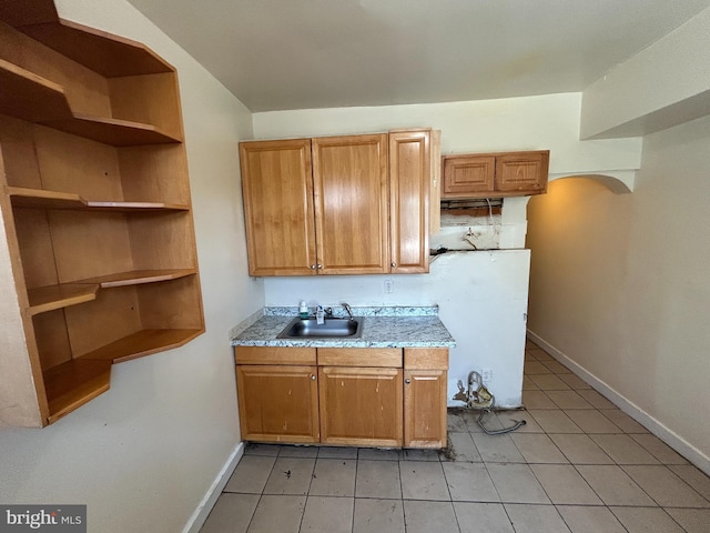 kitchen featuring a sink, baseboards, brown cabinetry, light tile patterned floors, and light stone countertops