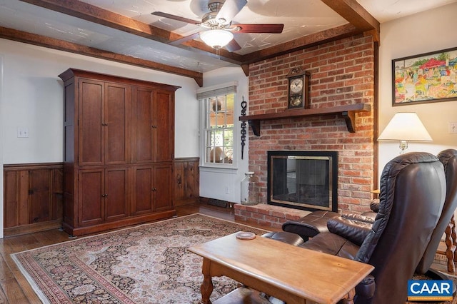 living room featuring wood-type flooring, a ceiling fan, a wainscoted wall, a brick fireplace, and beamed ceiling