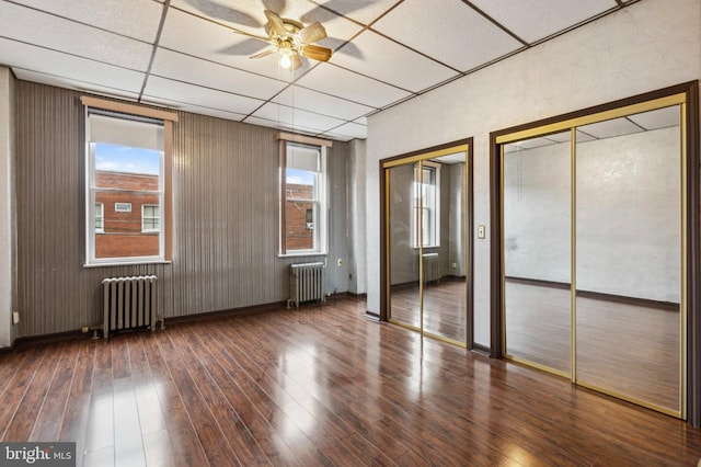 unfurnished bedroom featuring radiator heating unit, a drop ceiling, two closets, hardwood / wood-style flooring, and multiple windows