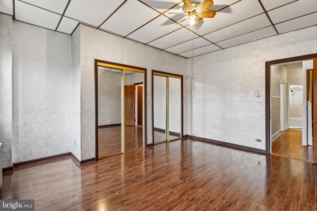unfurnished bedroom featuring two closets, wood finished floors, a paneled ceiling, baseboards, and a ceiling fan