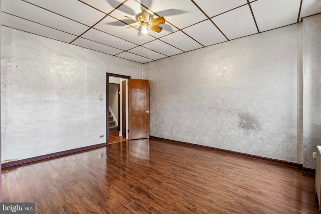 empty room featuring a drop ceiling, wood finished floors, a ceiling fan, baseboards, and stairway