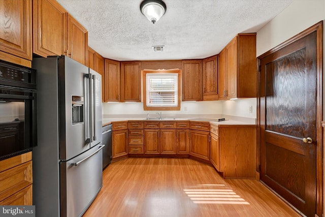 kitchen with appliances with stainless steel finishes, a textured ceiling, brown cabinetry, a sink, and light wood-style floors