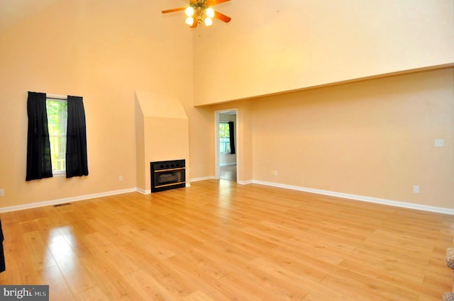 unfurnished living room featuring a ceiling fan, light wood-style flooring, a glass covered fireplace, a towering ceiling, and baseboards