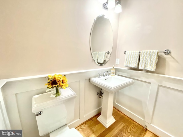 half bathroom with wood finished floors, a decorative wall, toilet, a wainscoted wall, and a sink