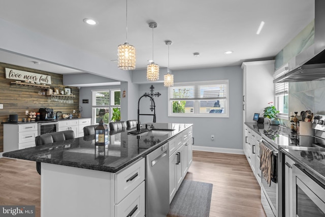 kitchen featuring stainless steel appliances, a sink, wall chimney range hood, a center island with sink, and white cabinets
