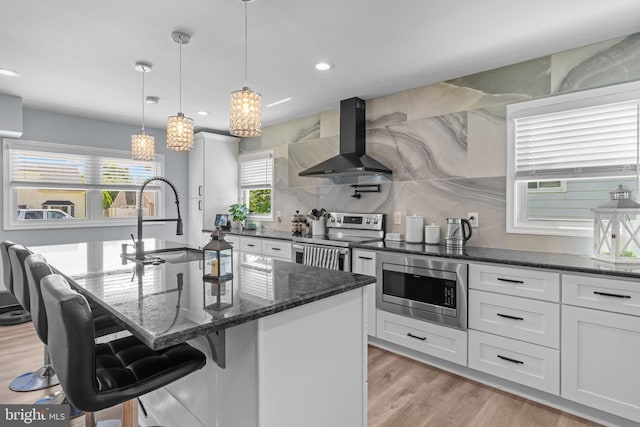 kitchen with wall chimney range hood, appliances with stainless steel finishes, a sink, dark stone counters, and white cabinets