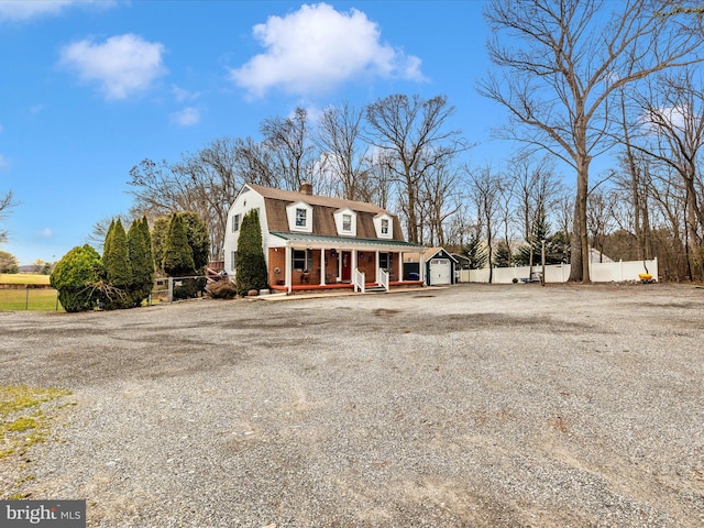 colonial inspired home with covered porch, a gambrel roof, fence, gravel driveway, and a chimney