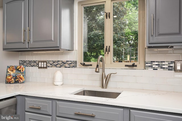 kitchen featuring gray cabinets, a sink, tasteful backsplash, light stone counters, and stainless steel dishwasher