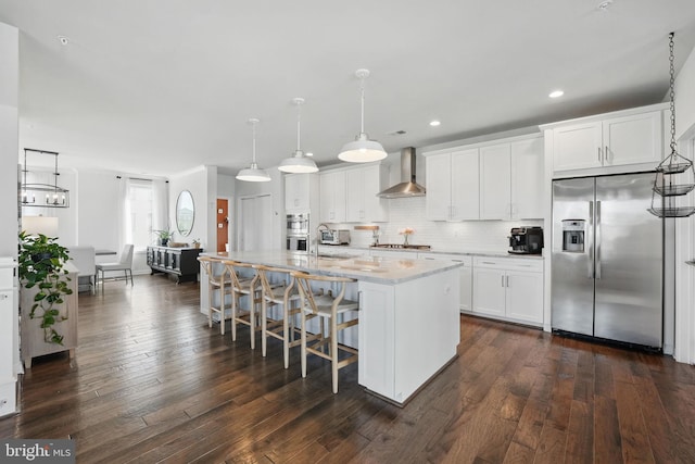 kitchen with stainless steel appliances, wall chimney exhaust hood, a kitchen island with sink, dark wood finished floors, and decorative backsplash