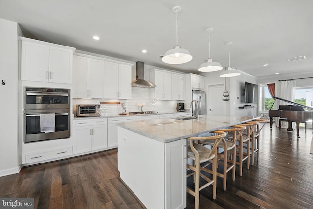 kitchen featuring wall chimney range hood, appliances with stainless steel finishes, a sink, a spacious island, and decorative backsplash