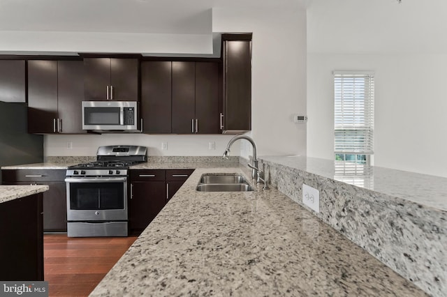 kitchen with stainless steel appliances, a sink, light stone counters, dark brown cabinetry, and dark wood-style flooring