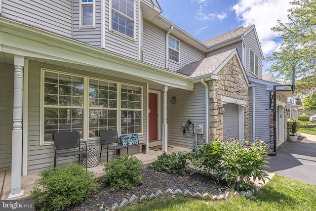 doorway to property with covered porch, a garage, stone siding, and a shingled roof
