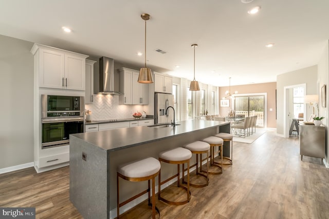 kitchen featuring stainless steel appliances, wall chimney exhaust hood, a kitchen island with sink, dark countertops, and baseboards