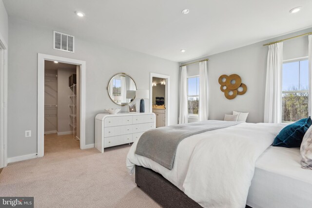 bedroom featuring a walk in closet, light colored carpet, recessed lighting, baseboards, and ensuite bath