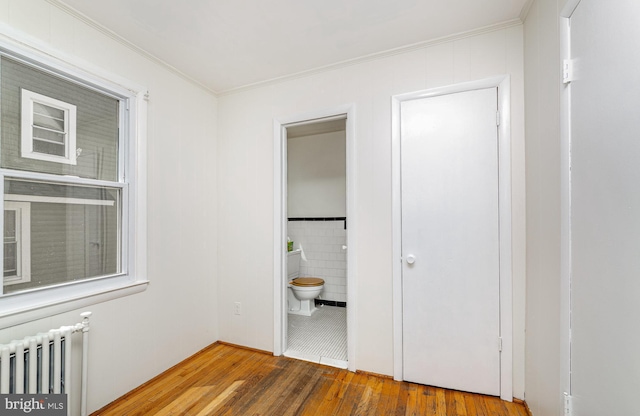 unfurnished bedroom with radiator, wood finished floors, crown molding, tile walls, and ensuite bath