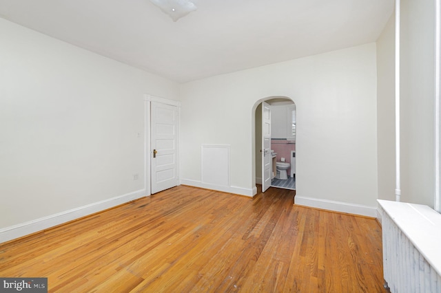 empty room with arched walkways, light wood-style flooring, baseboards, and radiator heating unit