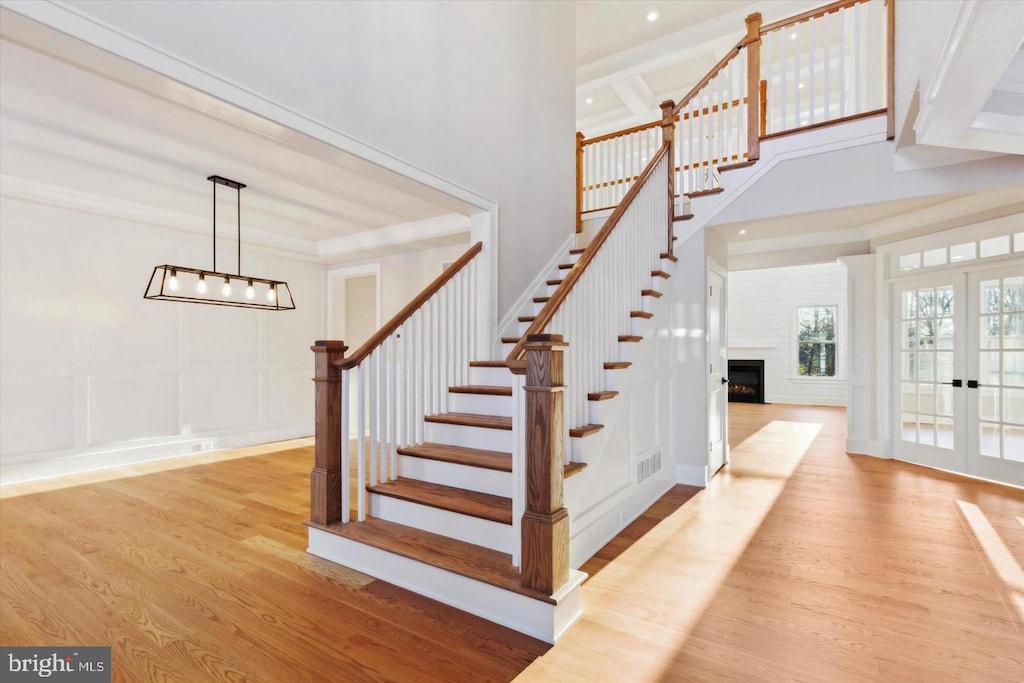stairway with a decorative wall, wood finished floors, french doors, a warm lit fireplace, and a chandelier