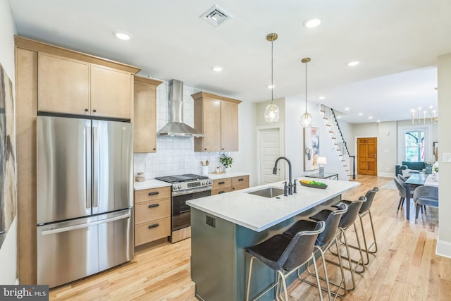 kitchen featuring stainless steel appliances, wall chimney range hood, a sink, light brown cabinets, and light wood-type flooring