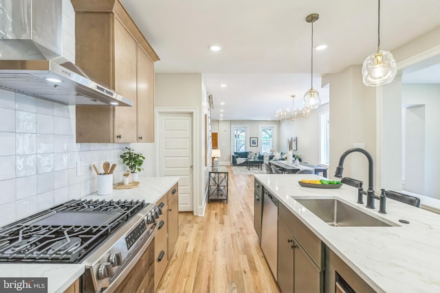 kitchen with wall chimney range hood, appliances with stainless steel finishes, a sink, light wood-type flooring, and light stone countertops