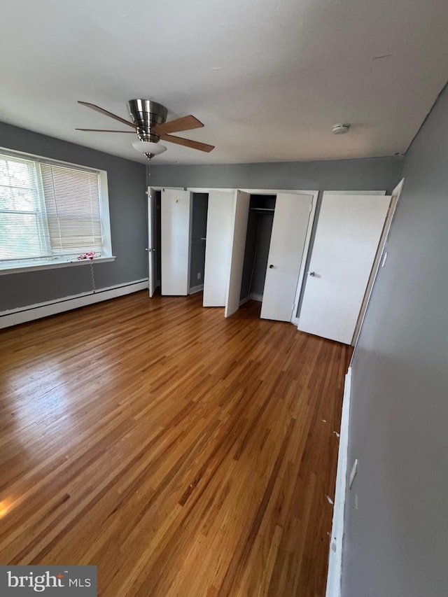 unfurnished bedroom featuring wood finished floors, a baseboard radiator, multiple closets, and a ceiling fan