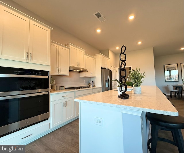 9181 Greenway Trl, Selbyville, DE 19975 - kitchen featuring appliances with stainless steel finishes, under cabinet range hood, white cabinets, dark wood finished floors, and a center island with sink