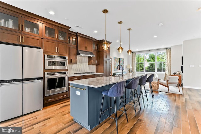 368 Star Tavern Ln, West Chester, PA 19382 - kitchen with appliances with stainless steel finishes, light wood-style floors, a center island with sink, under cabinet range hood, and light stone counters