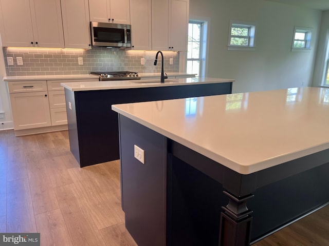 34232 Brenner Ln, Lewes, DE 19958 - kitchen featuring stainless steel microwave, an island with sink, light wood finished floors, decorative backsplash, and light countertops