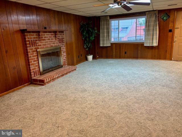 132 Popodickon Dr, Boyertown, PA 19512 - unfurnished living room featuring carpet floors, ceiling fan, wooden walls, a brick fireplace, and a drop ceiling