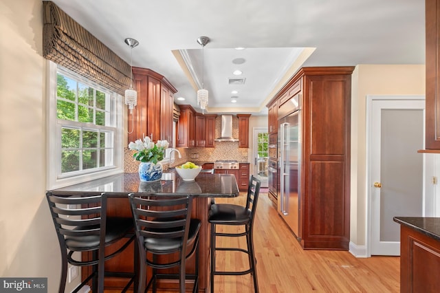 653 Broad Acres Rd, Penn Valley, PA 19072 - kitchen with wall chimney range hood, a raised ceiling, light wood-style floors, backsplash, and dark stone countertops