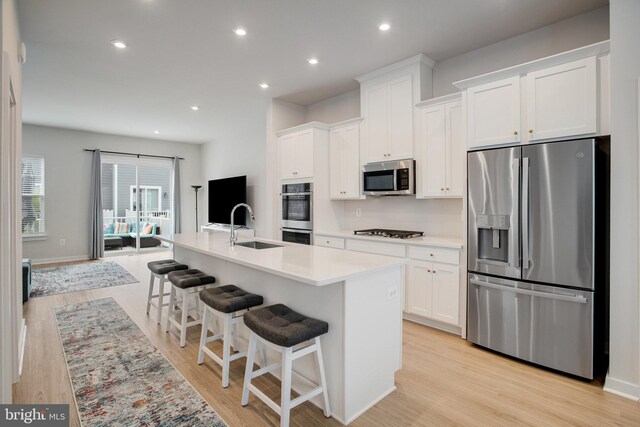 18351 Moss Garden Rd, Dumfries, VA 22026 - kitchen featuring appliances with stainless steel finishes, a kitchen island with sink, white cabinets, light wood-style floors, and a kitchen bar