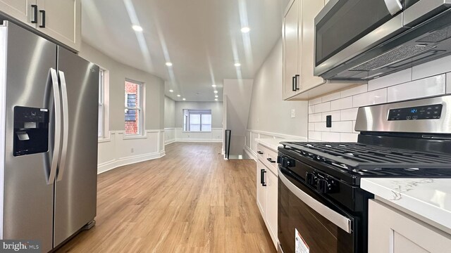 5841 Cedarhurst St, Philadelphia, PA 19143 - kitchen with appliances with stainless steel finishes, light wood-type flooring, backsplash, white cabinets, and a wainscoted wall
