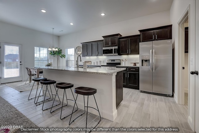 kitchen with light stone counters, appliances with stainless steel finishes, hanging light fixtures, a center island with sink, and an inviting chandelier