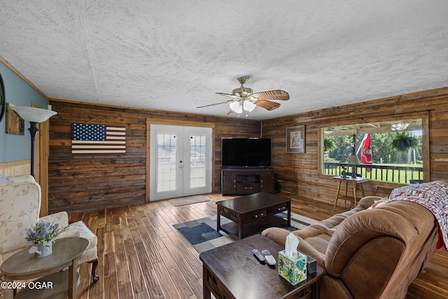 living room featuring french doors, a textured ceiling, wooden walls, wood-type flooring, and ceiling fan