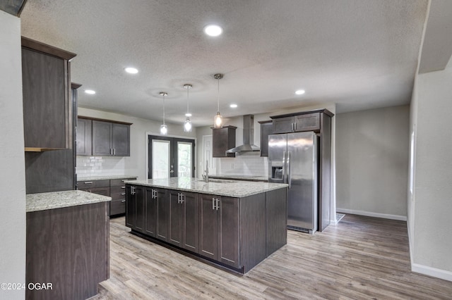 kitchen with stainless steel fridge, tasteful backsplash, wall chimney exhaust hood, light hardwood / wood-style flooring, and hanging light fixtures