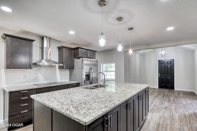 kitchen with a kitchen island with sink, sink, wall chimney range hood, decorative light fixtures, and stainless steel fridge with ice dispenser