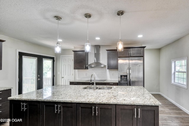 kitchen featuring stainless steel fridge, a center island with sink, wall chimney exhaust hood, and decorative light fixtures