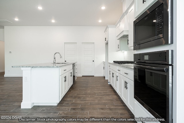 kitchen with built in microwave, white cabinetry, light stone counters, a center island with sink, and oven